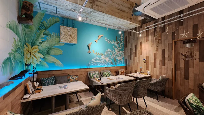 Comedor con arte mural tropical y sillas de mimbre en Hawaiian Café Kapuka en American Village en Chatan
